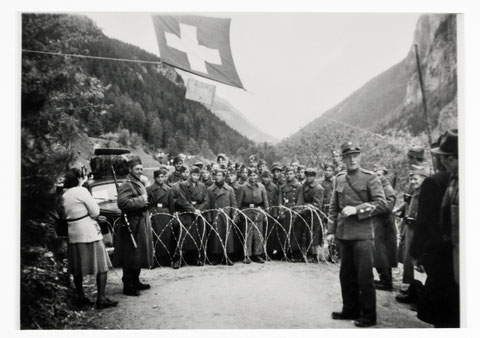 Menschen drängen sich hinter einer Stacheldrahtabsperrung. Im Vordergrund einige Soldaten, im Hintergrund Berge. Über allem weht eine Schweizer Fahne.